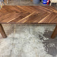 A rectangular wooden dining table with a herringbone pattern tabletop and block legs, photographed in a workshop or garage setting on a concrete floor.
