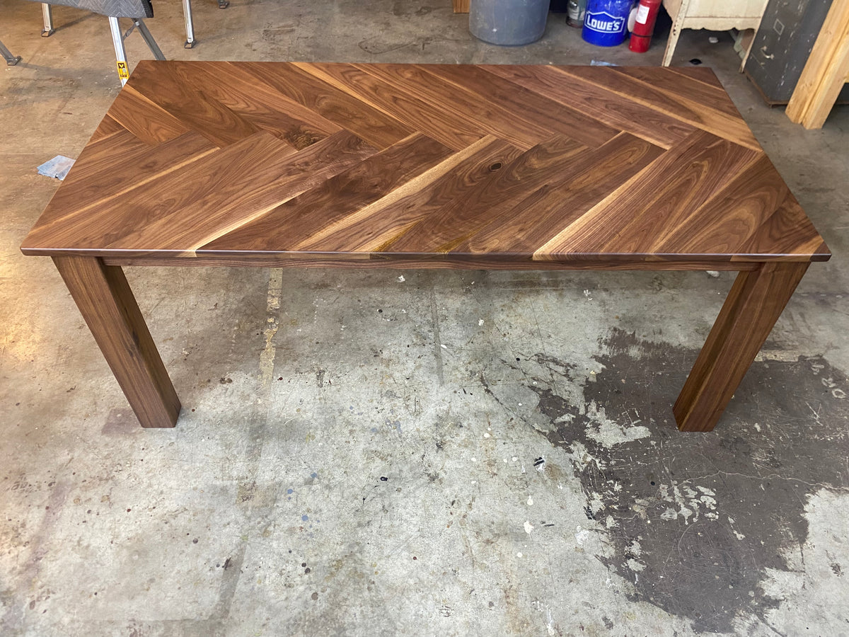 A rectangular wooden dining table with a herringbone pattern tabletop and block legs, photographed in a workshop or garage setting on a concrete floor.