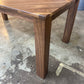A close-up of a wooden dining table built with walnut, showing the tabletop's herringbone pattern and one of its sturdy legs, set on a concrete workshop floor.