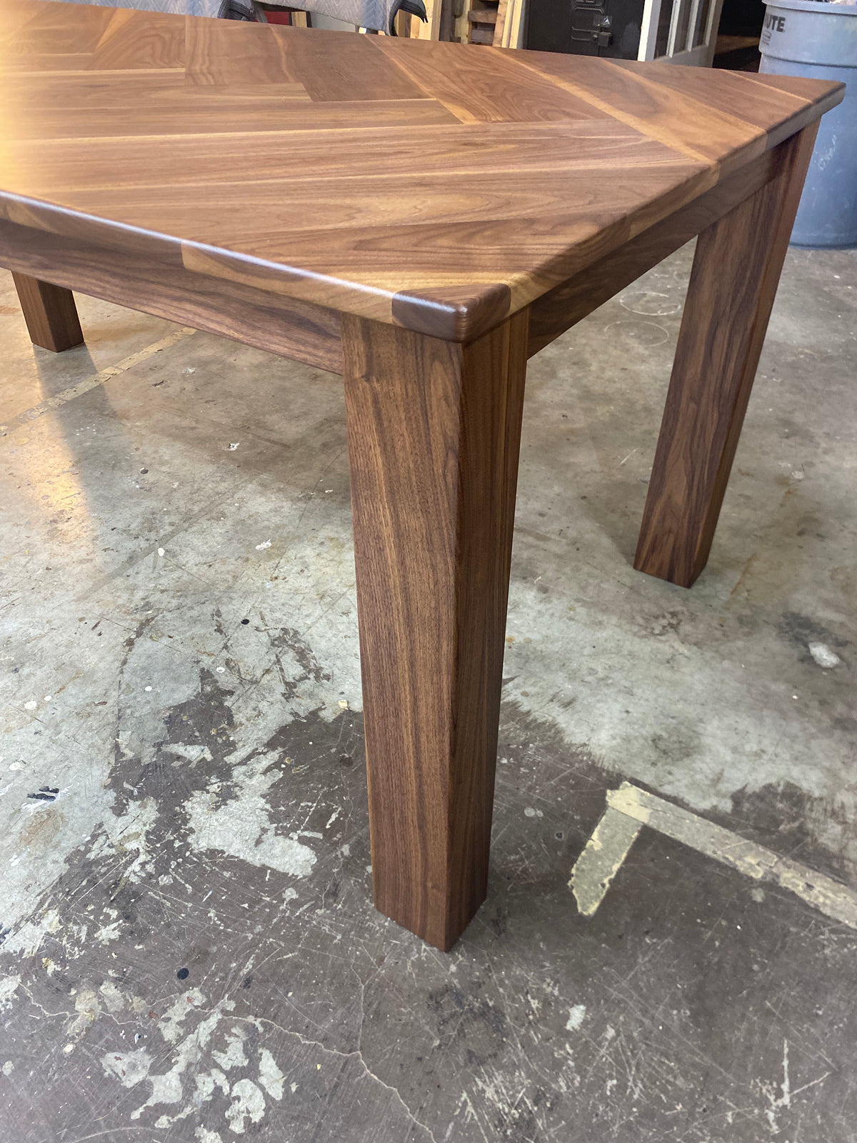 A close-up of a wooden dining table built with walnut, showing the tabletop's herringbone pattern and one of its sturdy legs, set on a concrete workshop floor.