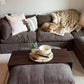 A cozy living room scene featuring a dark brown sectional sofa with a small golden dog sleeping on the seat cushion, wrapped in a chunky knit beige throw blanket. In front of the sofa is a dark wood coffee table with a matching ottoman, styled with a small potted plant and a white bowl on a tray.