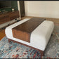 An oversized wood ottoman table with a dark walnut stained tray on top of an ottoman, placed in a room with a wooden floor and a multicolored area rug.