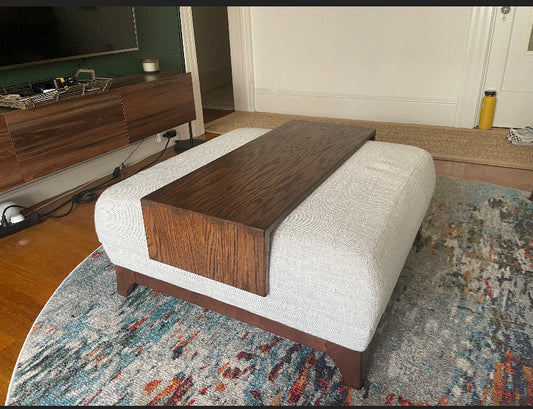 An oversized wood ottoman table with a dark walnut stained tray on top of an ottoman, placed in a room with a wooden floor and a multicolored area rug.