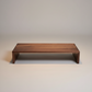 A minimalist wooden low table or bench with a warm medium-brown wood grain finish, photographed against a seamless light gray studio background.