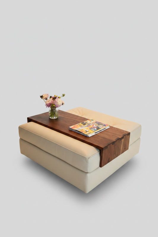 A modern beige upholstered ottoman or coffee table with a dark wood top panel, styled with a small vase of flowers and a magazine, on a solid light gray background.